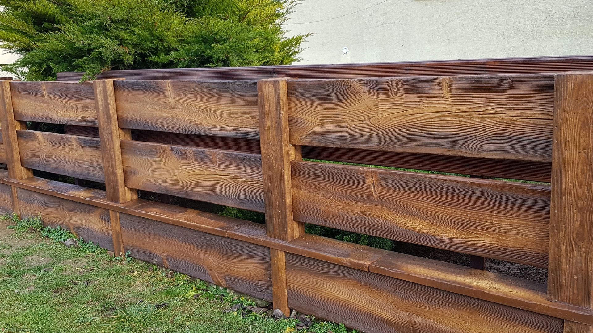 Betonowe ogrodzenie imitujące drewno, z widoczną strukturą słojów, z zielenią w tle.