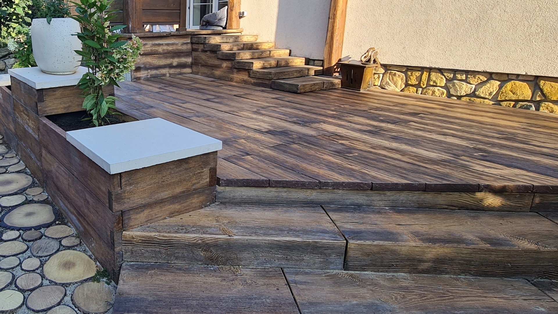 Betonowy taras z desek i schody w rustykalnym stylu, uzupełnione donicami z roślinami, prowadzące do wejścia domu z elewacją wykończoną kamieniem.