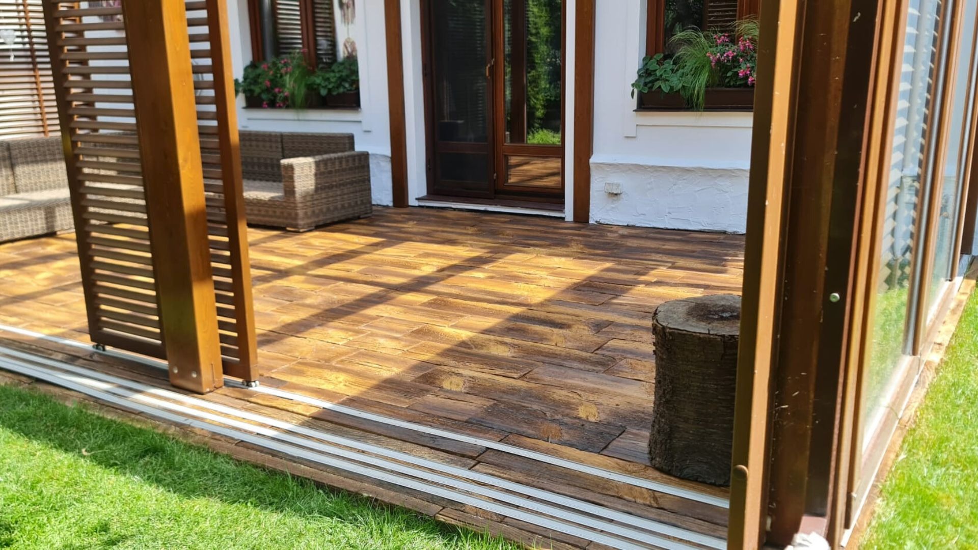 Zadaszony taras z drewnopodobną posadzką, przesuwnymi panelami z lamelami i widokiem na dom z oknami w kwiatach.