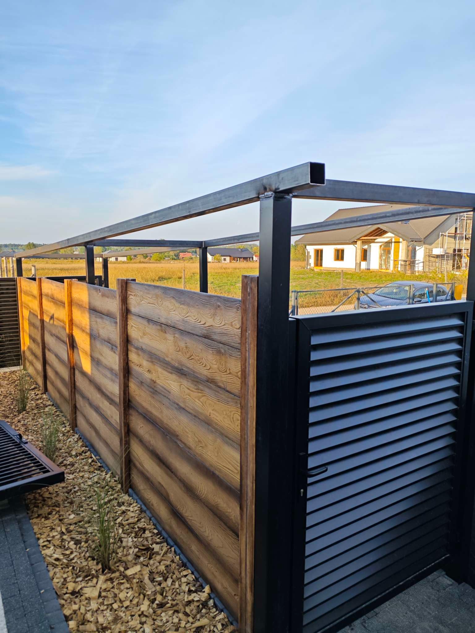 Nowoczesne ogrodzenie z elementami drewnopodobnymi i metalową bramą lamelową w kolorze czarnym, z widoczną konstrukcją pergoli i zabudową na posesji w tle.