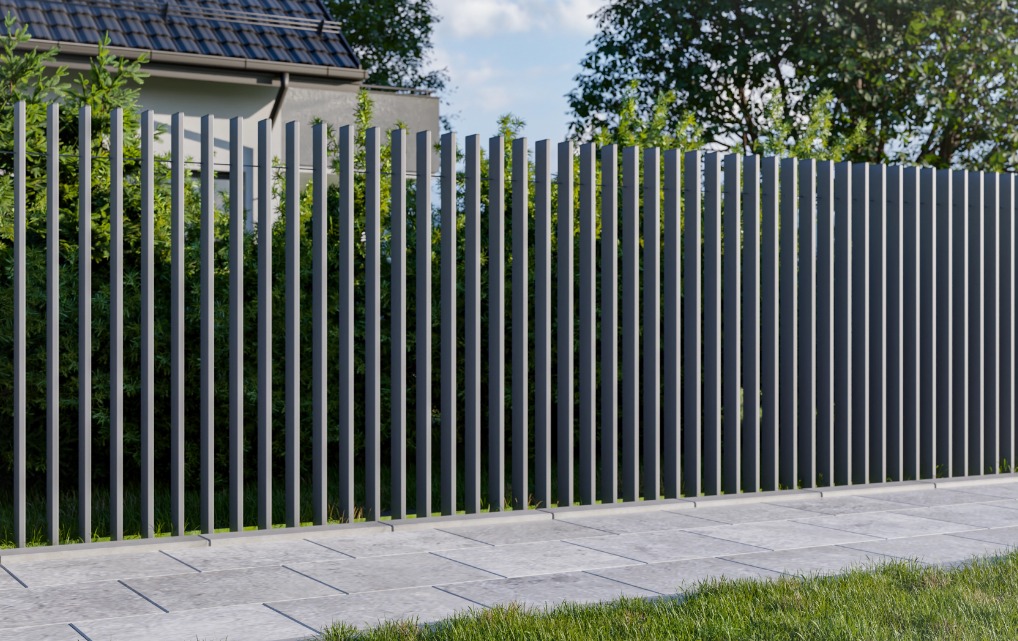 Nowoczesne ogrodzenie metalowe z pionowych, wąskich profili w kolorze grafitowym, oddzielające posesję od chodnika z szarych płyt, z zielenią w tle.