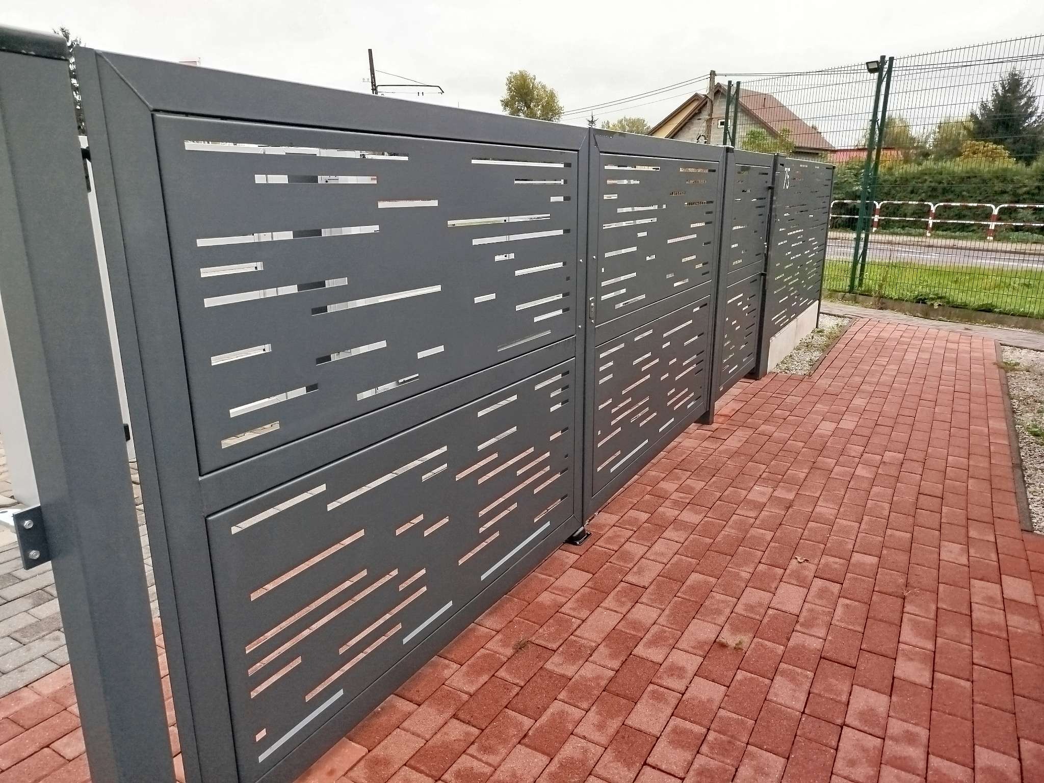 Nowoczesne, szare przęsła ogrodzeniowe z poziomymi, prostokątnymi wycięciami, ustawione na kostce brukowej w kolorze ceglastym, widok z boku.