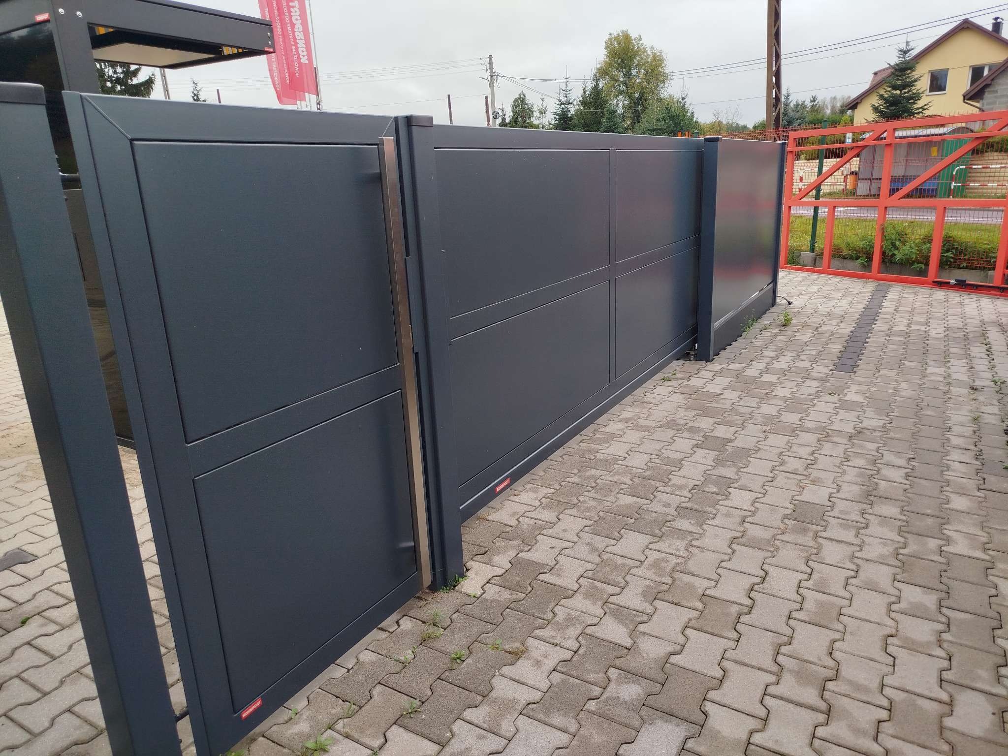 Nowoczesna, przesuwna brama w kolorze antracytowym, z widocznymi panelami i subtelnymi, pionowymi uchwytami, zamontowana na posesji z kostką brukową, z częściowo otwartym skrzydłem, ukazującym...