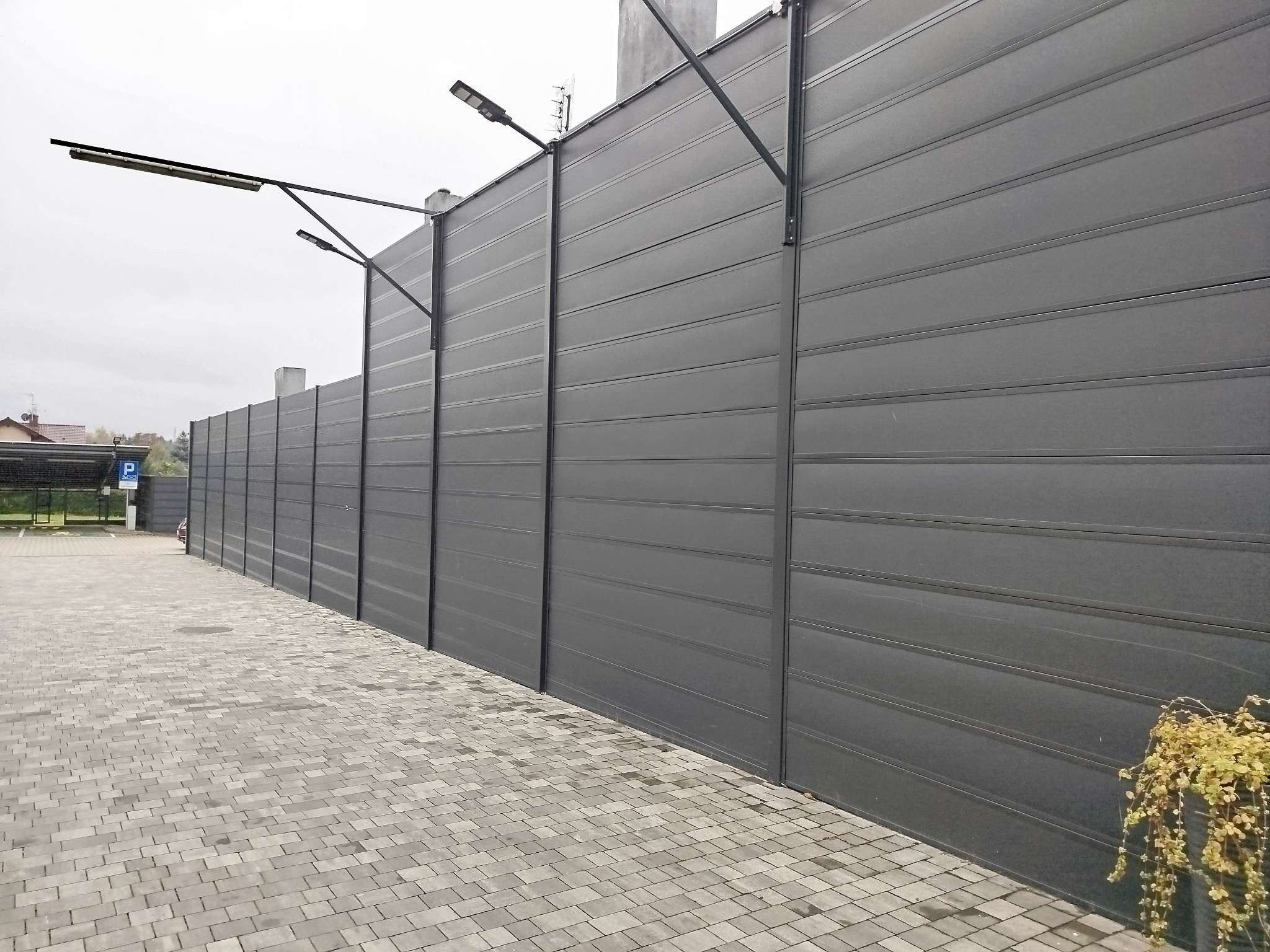 Długie, szare ogrodzenie panelowe z oświetleniem LED wzdłuż chodnika z kostki brukowej, widok z perspektywy przechodnia.