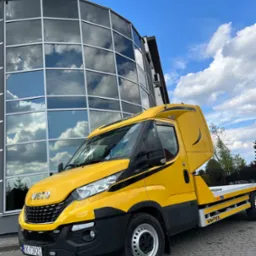 Żółta laweta Iveco z tablicą rejestracyjną KK 7392G zaparkowana przed nowoczesnym budynkiem z lustrzanymi oknami odbijającymi błękitne niebo z chmurami.