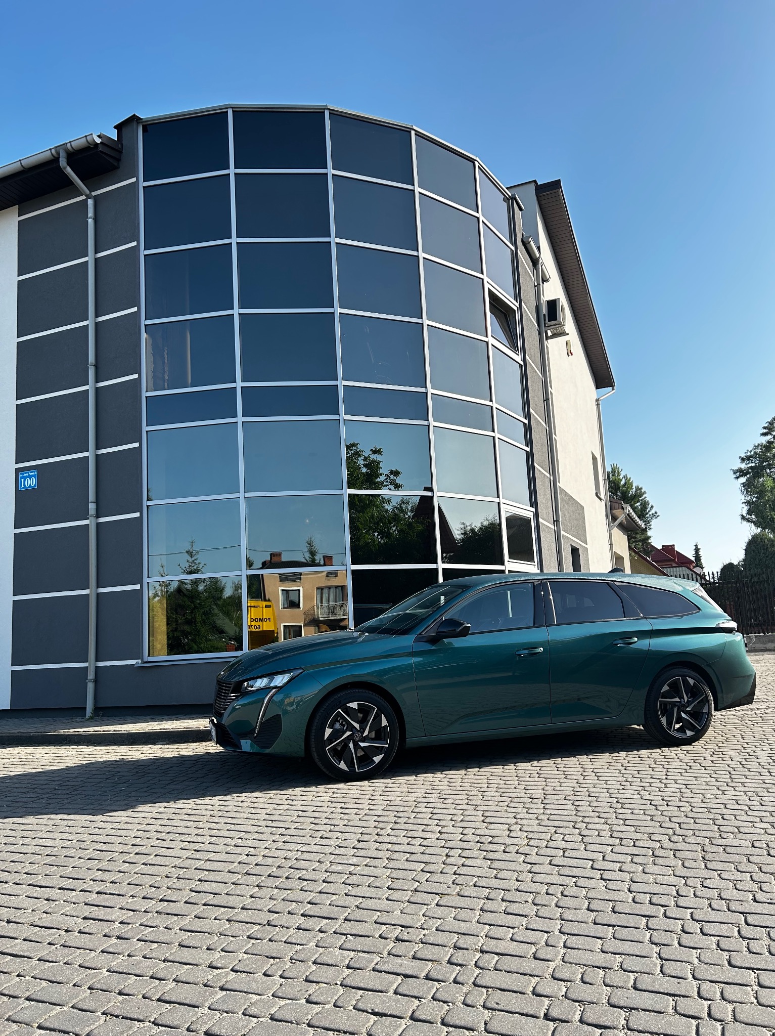 Zielony samochód kombi Peugeot parkuje na brukowanej ulicy przed nowoczesnym budynkiem biurowym z dużą szklaną fasadą, odbijającą otoczenie.