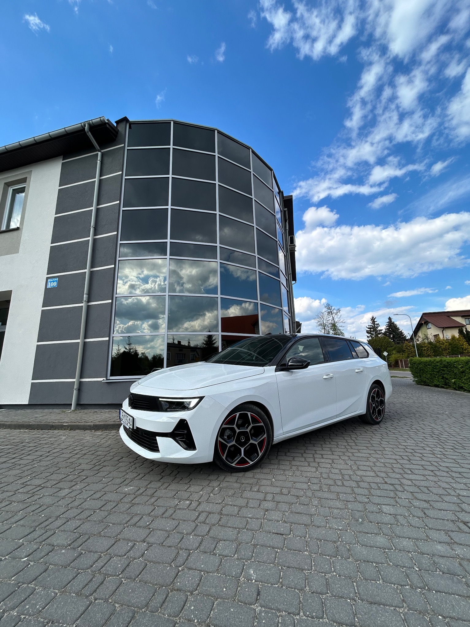 Biały samochód kombi z czarnym dachem i czerwonymi akcentami na felgach, zaparkowany na brukowanym podjeździe przed nowoczesnym budynkiem z dużymi oknami, w których odbija się błękitne niebo...