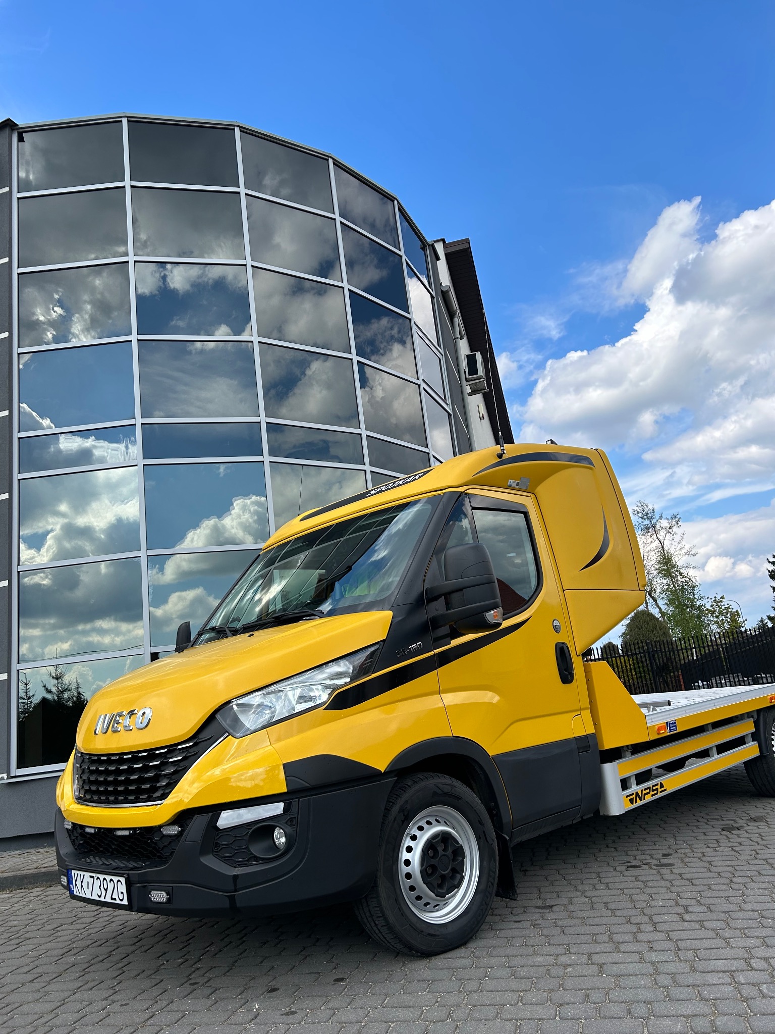 Żółta laweta Iveco z tablicą rejestracyjną KK 7392G zaparkowana przed nowoczesnym budynkiem z lustrzanymi oknami odbijającymi błękitne niebo z chmurami.