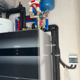 Nowoczesny piec grzewczy marki Buderus z podłączonymi rurami instalacji CO w kolorach czarnym, czerwonym i niebieskim, widoczny także zbiornik wyrównawczy oraz skrzynka elektryczna na ścianie.