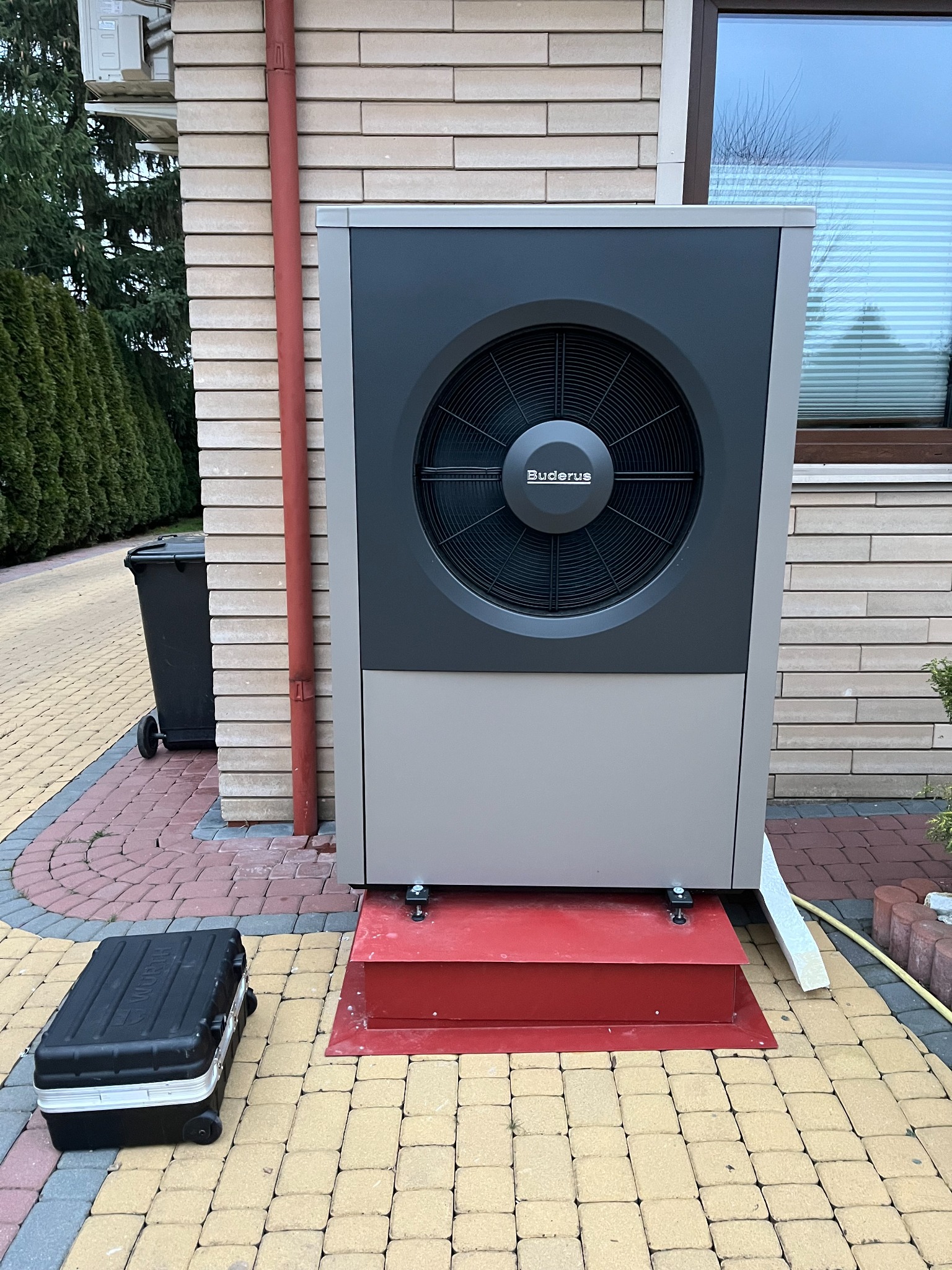 Zewnętrzna jednostka pompy ciepła marki Buderus zamontowana na czerwonym podeście, obok skrzynka narzędziowa na kostce brukowej.