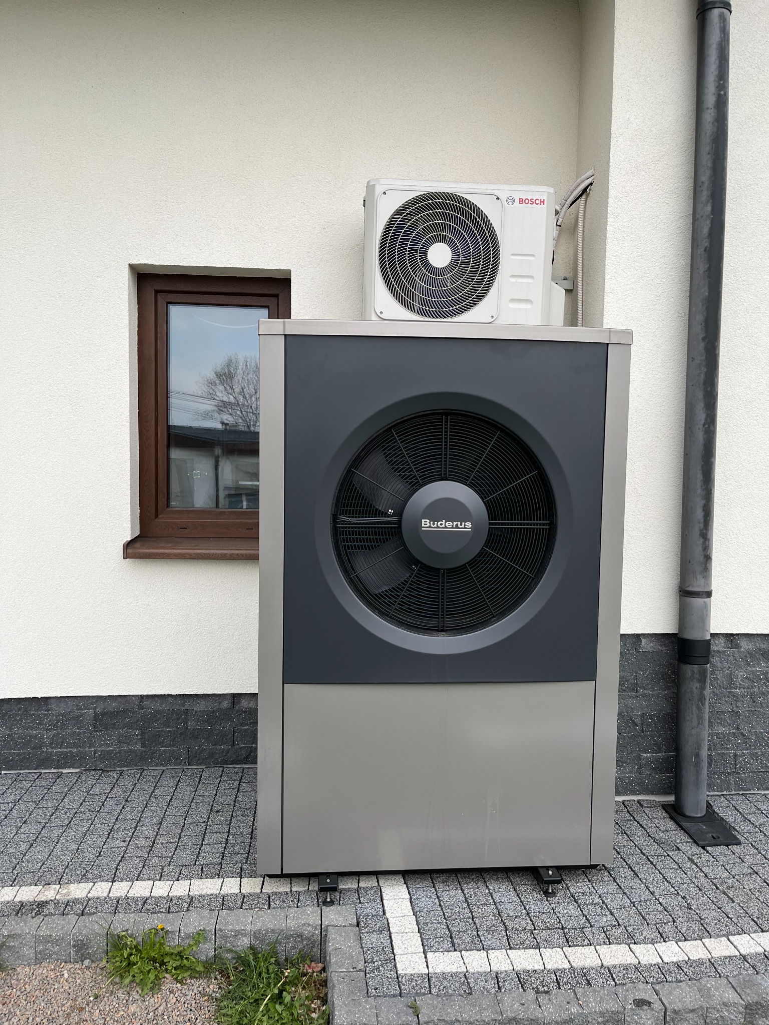 Zewnętrzna instalacja dwuczęściowej pompy ciepła Buderus i Bosch zamontowana przy ścianie domu z widocznym oknem i rurą spustową