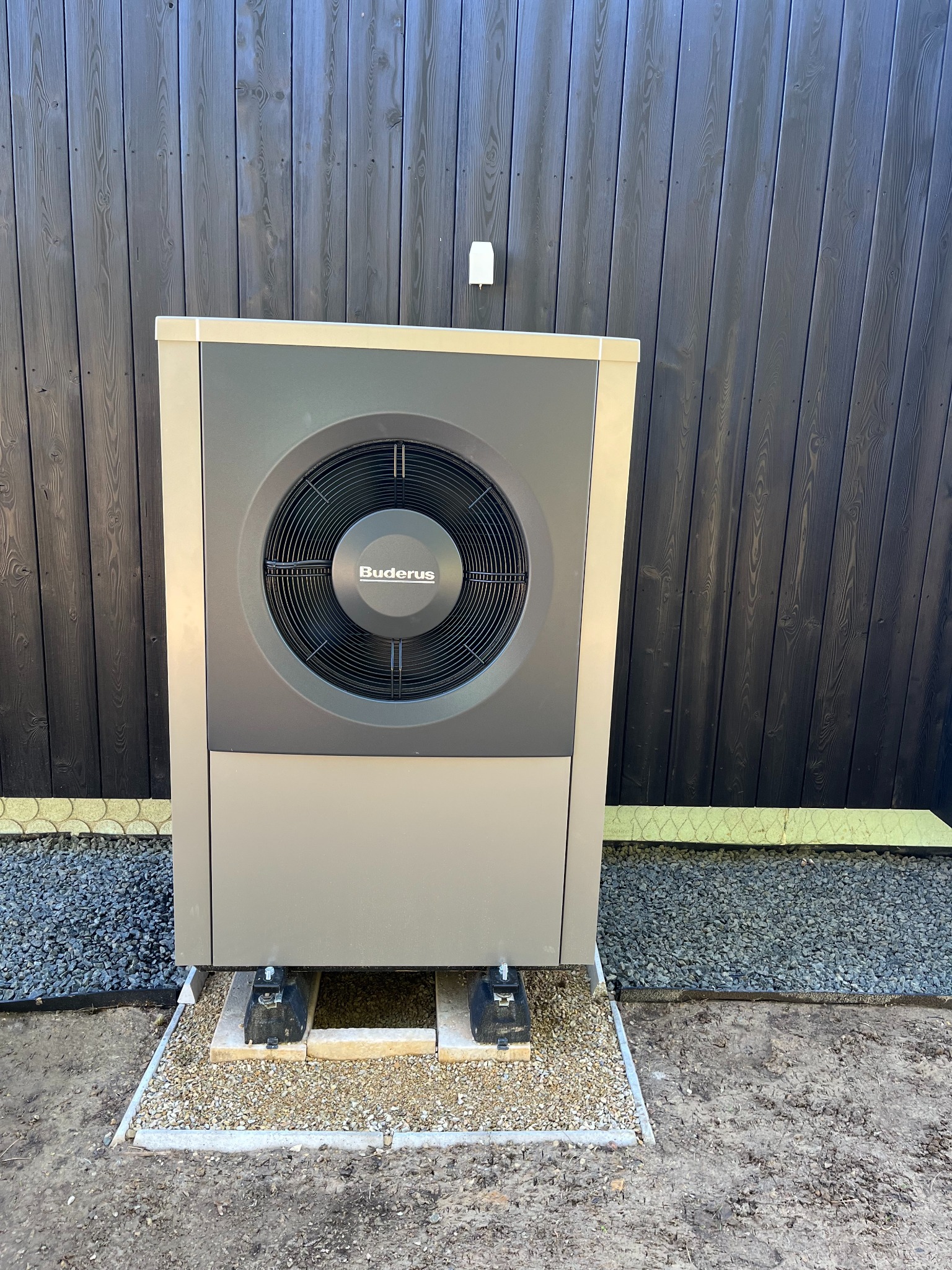 Szara pompa ciepła Buderus zainstalowana przy ciemnej, drewnianej ścianie budynku, umieszczona na betonowej podstawie wypełnionej drobnym żwirem.