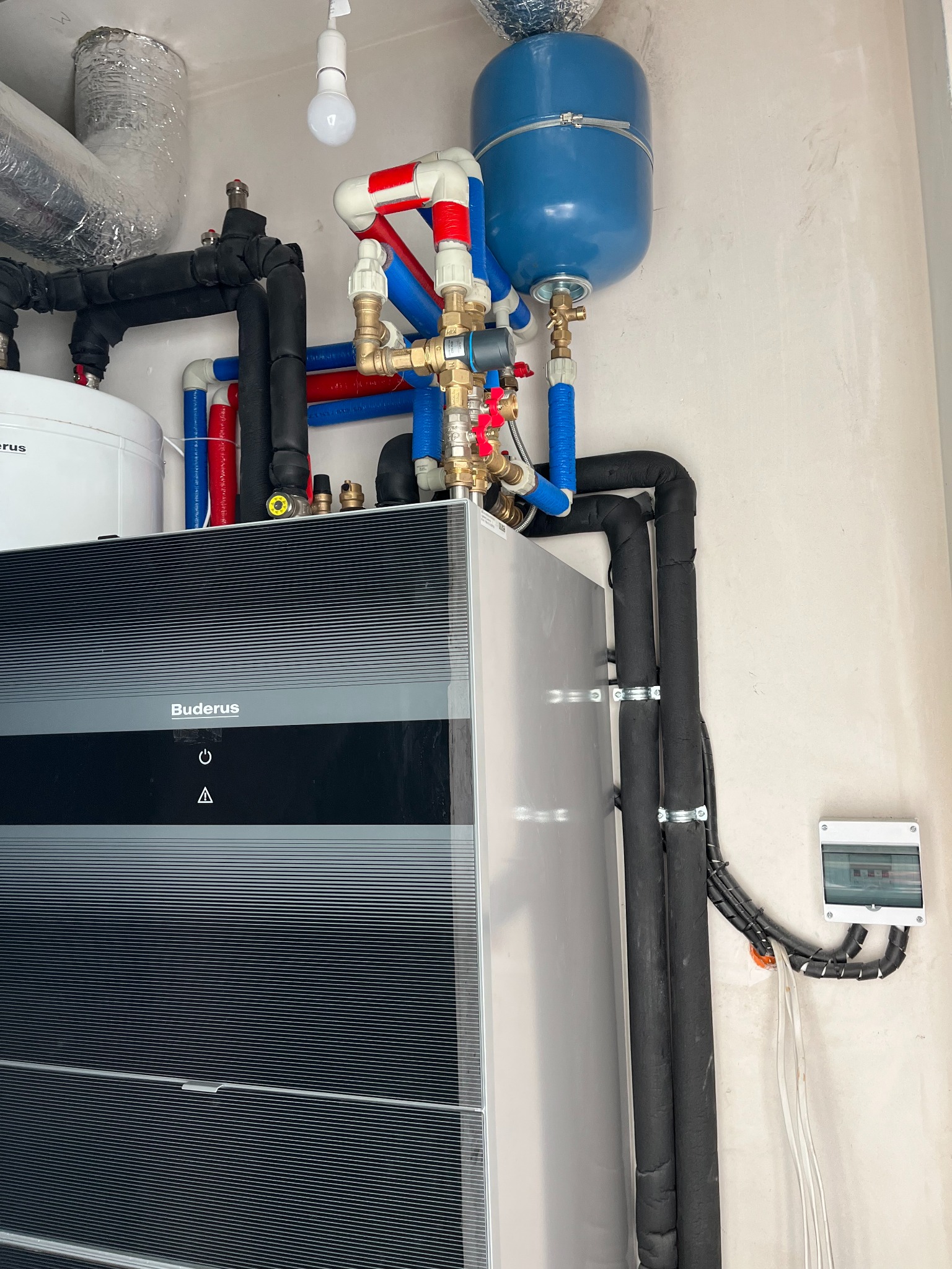 Nowoczesny piec grzewczy marki Buderus z podłączonymi rurami instalacji CO w kolorach czarnym, czerwonym i niebieskim, widoczny także zbiornik wyrównawczy oraz skrzynka elektryczna na ścianie.