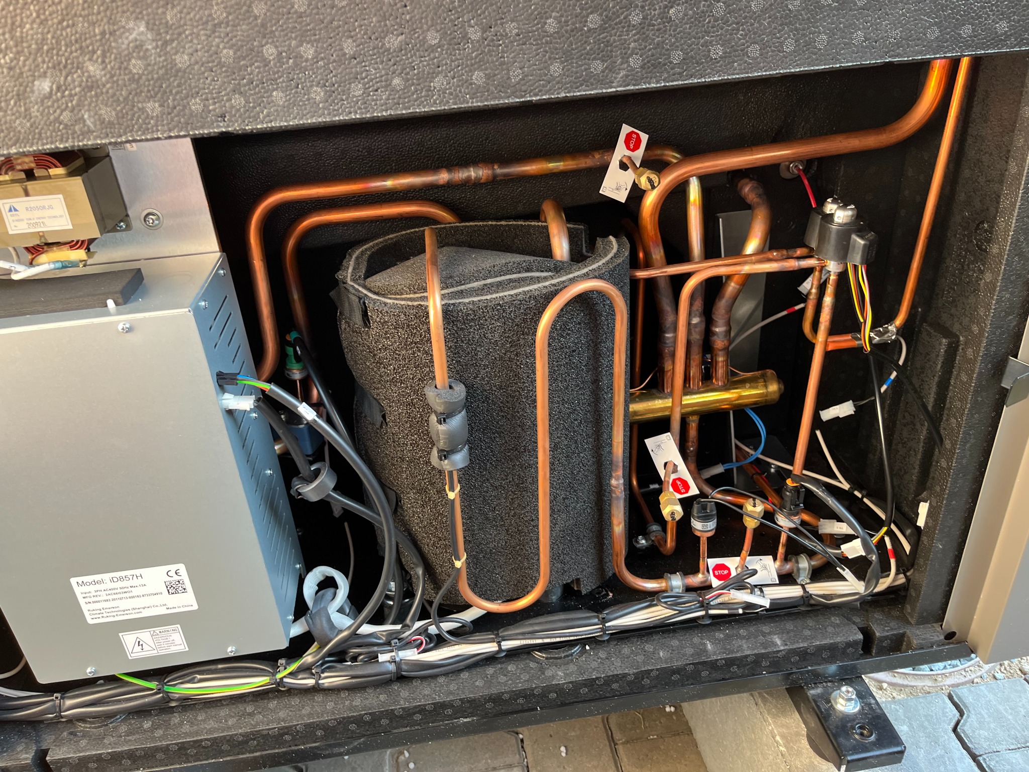 Wnętrze urządzenia grzewczego z widocznymi miedzianymi rurami, izolowanym zbiornikiem, elementami elektrycznymi i tabliczką znamionową modelu iD857H. Widoczne etykiety ostrzegawcze i oznaczenia...