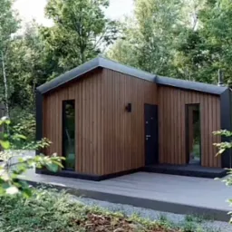 Nowoczesny, drewniany domek letniskowy z płaskim dachem, otoczony zielenią, z widocznymi pionowymi panelami elewacyjnymi i minimalistycznym oświetleniem zewnętrznym.