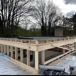 Jan Kaluza - Wznoszenie drewnianej konstrukcji domu w stanie surowym otwartym, z widoczną izolacją fundamentów i czarną papą na dachu, na tle drzew i pochmurnego nieba.