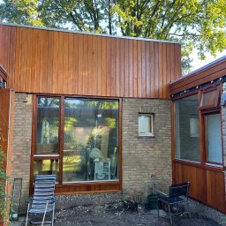 Jan Kaluza - Elewacja domu z drewnianą okładziną, widoczne okna w drewnianych ramach, ceglana ściana, krzesła ogrodowe i elementy wyposażenia wnętrza w trakcie prac wykończeniowych.