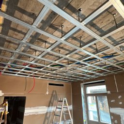 Jan Kaluza - Montaż metalowej konstrukcji sufitu podwieszanego z widocznym ociepleniem stropu oraz instalacją elektryczną, drabina oparta o ścianę z płyt kartonowo-gipsowych, okno w tle.
