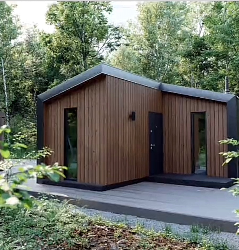 Nowoczesny, drewniany domek letniskowy z płaskim dachem, otoczony zielenią, z widocznymi pionowymi panelami elewacyjnymi i minimalistycznym oświetleniem zewnętrznym.