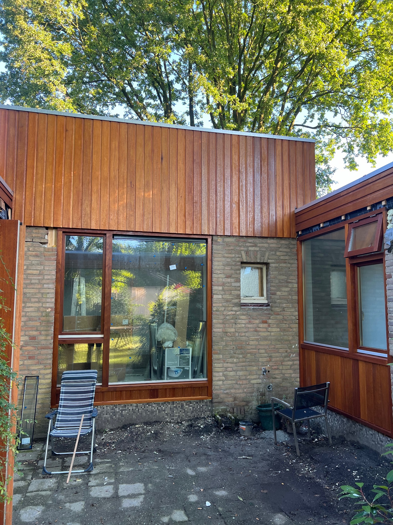 Elewacja domu z drewnianą okładziną, widoczne okna w drewnianych ramach, ceglana ściana, krzesła ogrodowe i elementy wyposażenia wnętrza w trakcie prac wykończeniowych.