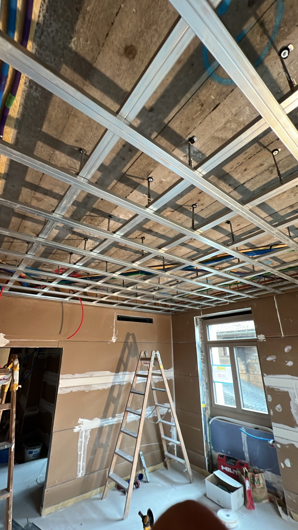 Montaż metalowej konstrukcji sufitu podwieszanego z widocznym ociepleniem stropu oraz instalacją elektryczną, drabina oparta o ścianę z płyt kartonowo-gipsowych, okno w tle.