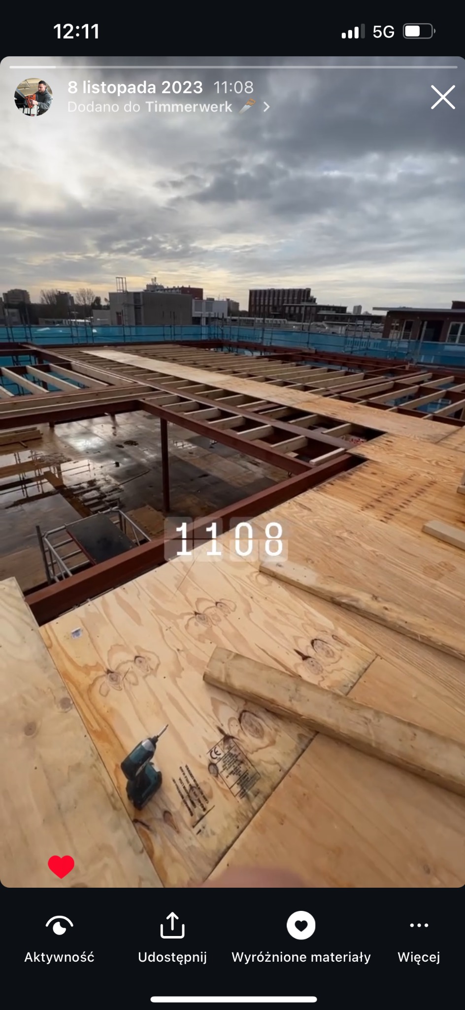 Konstrukcja drewniana dachu płaskiego w trakcie budowy, widoczne płyty OSB, metalowe belki, narzędzie - wkrętarka akumulatorowa leżąca na płycie, tło stanowi zabudowa miejska pod pochmurnym niebem.