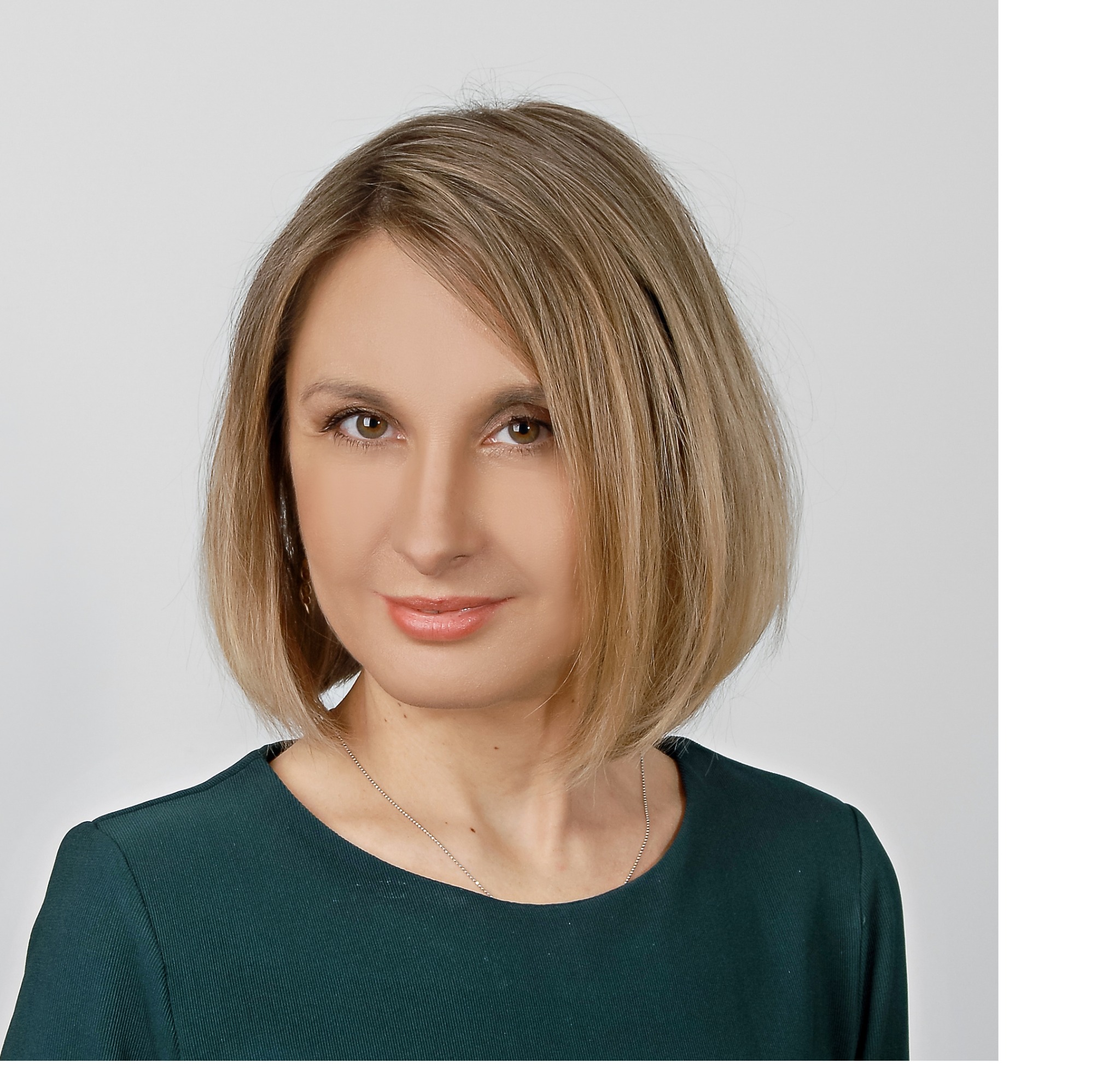 Portret kobiety z krótkimi blond włosami, ubranej w ciemnozieloną sukienkę, na jasnym tle, z delikatnym uśmiechem.