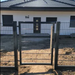 Nowoczesna, grafitowa furtka panelowa z widokiem na dom jednorodzinny z ciemną dachówką i jasną elewacją, na tle błękitnego nieba.