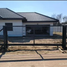 Nowoczesna, przesuwna brama wjazdowa z metalową siatką na tle białego domu z ciemną dachówką. Widoczne elementy montażowe i piasek. Słoneczny dzień.