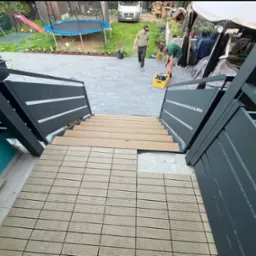 Montaż drewnianej podłogi tarasowej na schodach z metalową balustradą w ogrodzie. Widoczny etap prac, narzędzia i osoby w tle. Kąt ujęcia z góry.