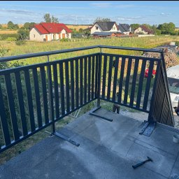 KRYCMET YEVHEN CHEKERLAN - Montaż metalowej balustrady balkonowej z pionowymi szczeblami, widok na okoliczne domy i pola w słoneczny dzień. Etap mocowania balustrady do podłoża.