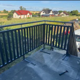 Montaż metalowej balustrady balkonowej z pionowymi szczeblami, widok na okoliczne domy i pola w słoneczny dzień. Etap mocowania balustrady do podłoża.