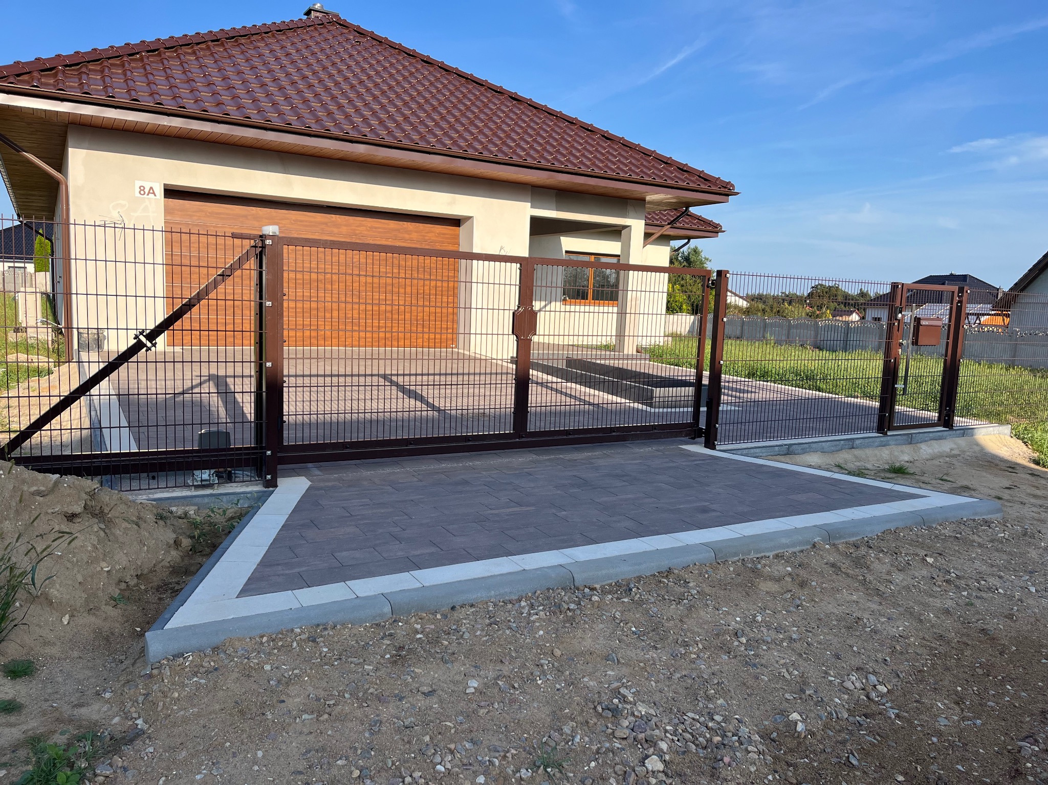 Nowoczesna posesja z brązową bramą przesuwną i dopasowanym kolorystycznie ogrodzeniem panelowym, kostka brukowa ułożona na podjeździe, dom z brązowym dachem w tle.