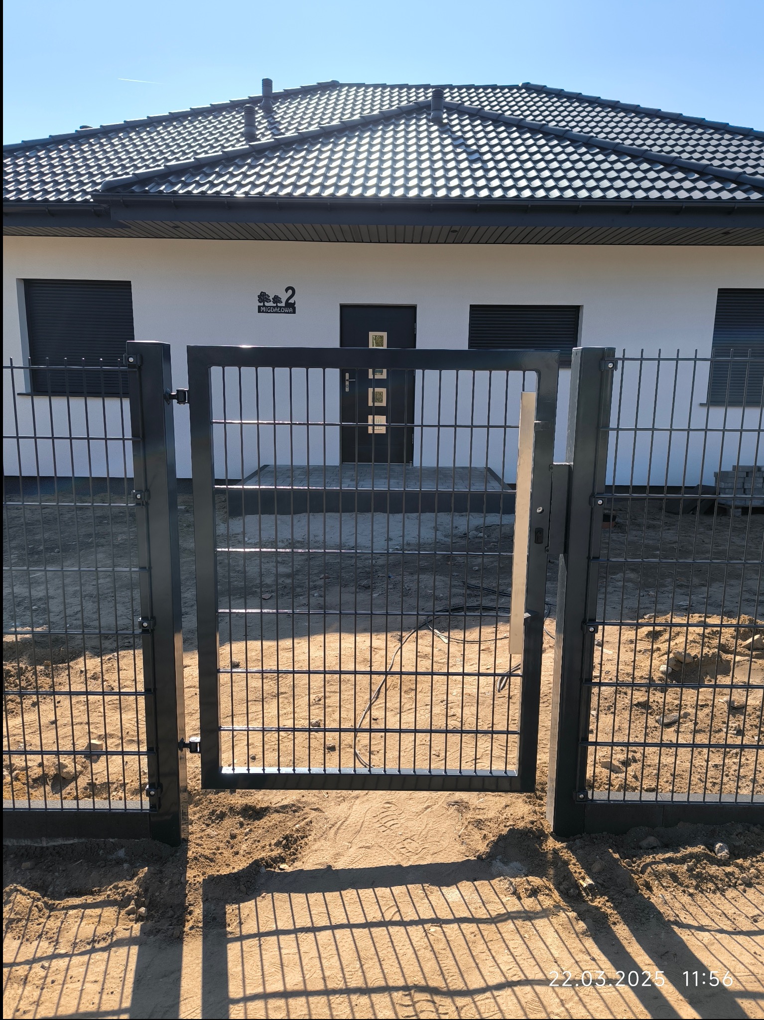 Nowoczesna, grafitowa furtka panelowa z widokiem na dom jednorodzinny z ciemną dachówką i jasną elewacją, na tle błękitnego nieba.