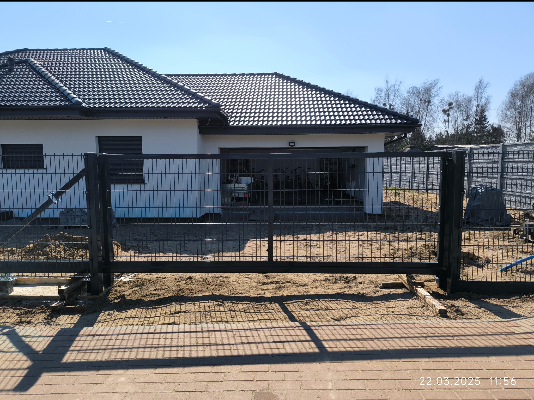 Nowoczesna, przesuwna brama wjazdowa z metalową siatką na tle białego domu z ciemną dachówką. Widoczne elementy montażowe i piasek. Słoneczny dzień.