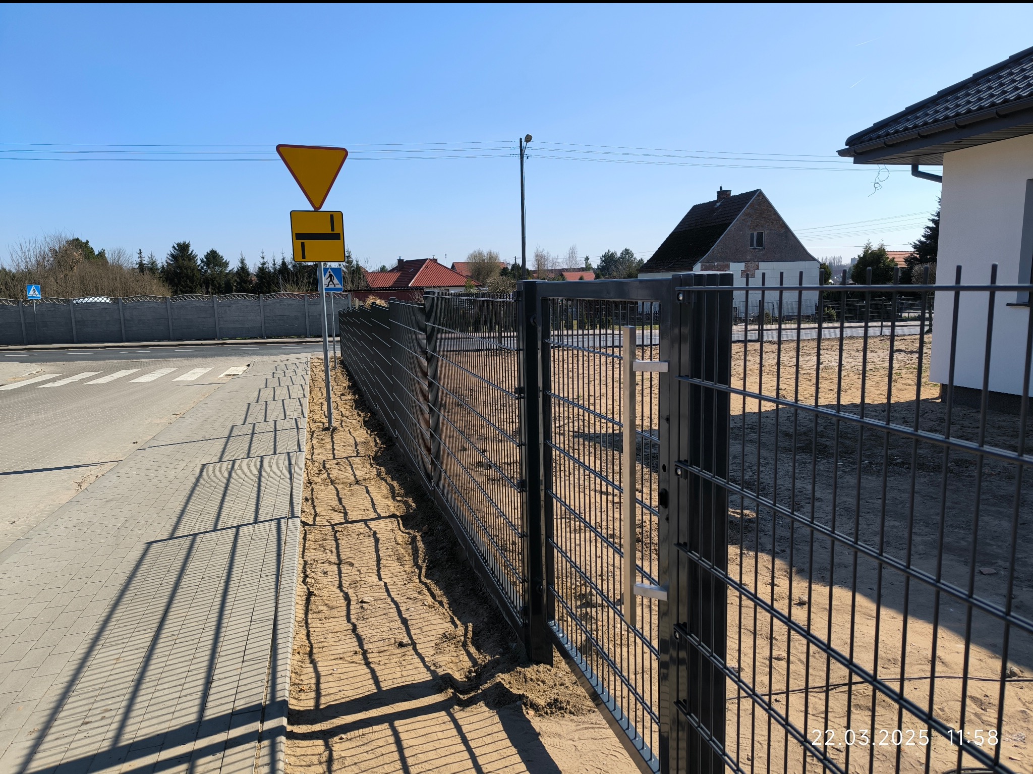 Nowoczesne ogrodzenie panelowe w kolorze grafitowym, oddzielające posesję od chodnika. W tle domy i znaki drogowe. Cień ogrodzenia na piasku.
