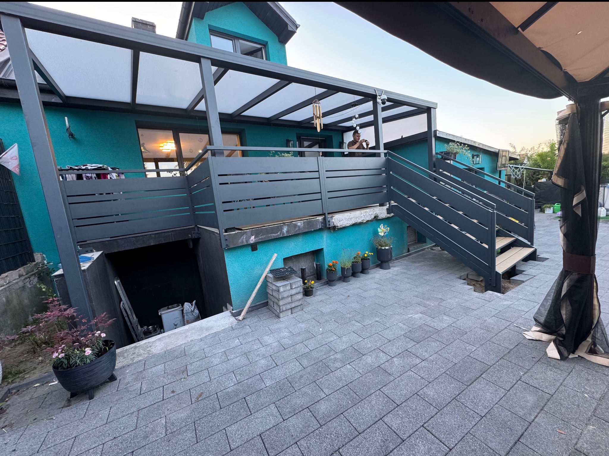 Nowoczesna pergola z metalową konstrukcją i zadaszeniem z poliwęglanu nad balkonem domu o turkusowej elewacji, z metalową balustradą i schodami.