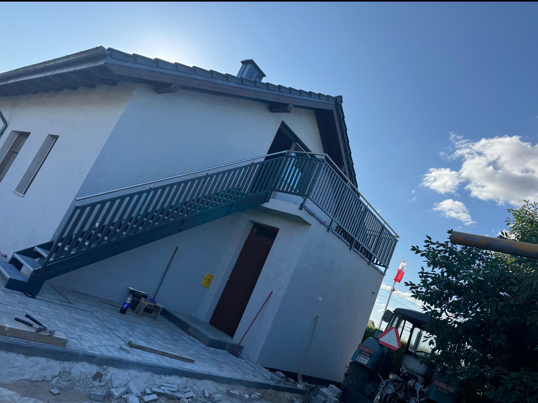 Metalowe schody zewnętrzne z balustradą prowadzące na balkon domu jednorodzinnego w trakcie wykańczania. Widoczny ciągnik i narzędzia.