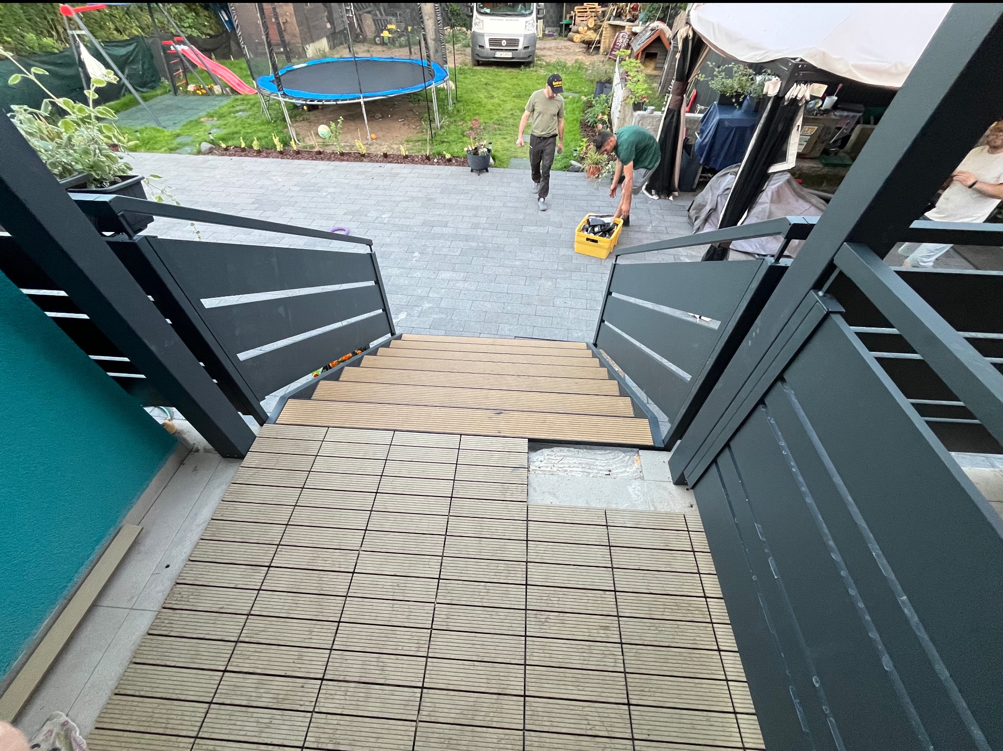 Montaż drewnianej podłogi tarasowej na schodach z metalową balustradą w ogrodzie. Widoczny etap prac, narzędzia i osoby w tle. Kąt ujęcia z góry.