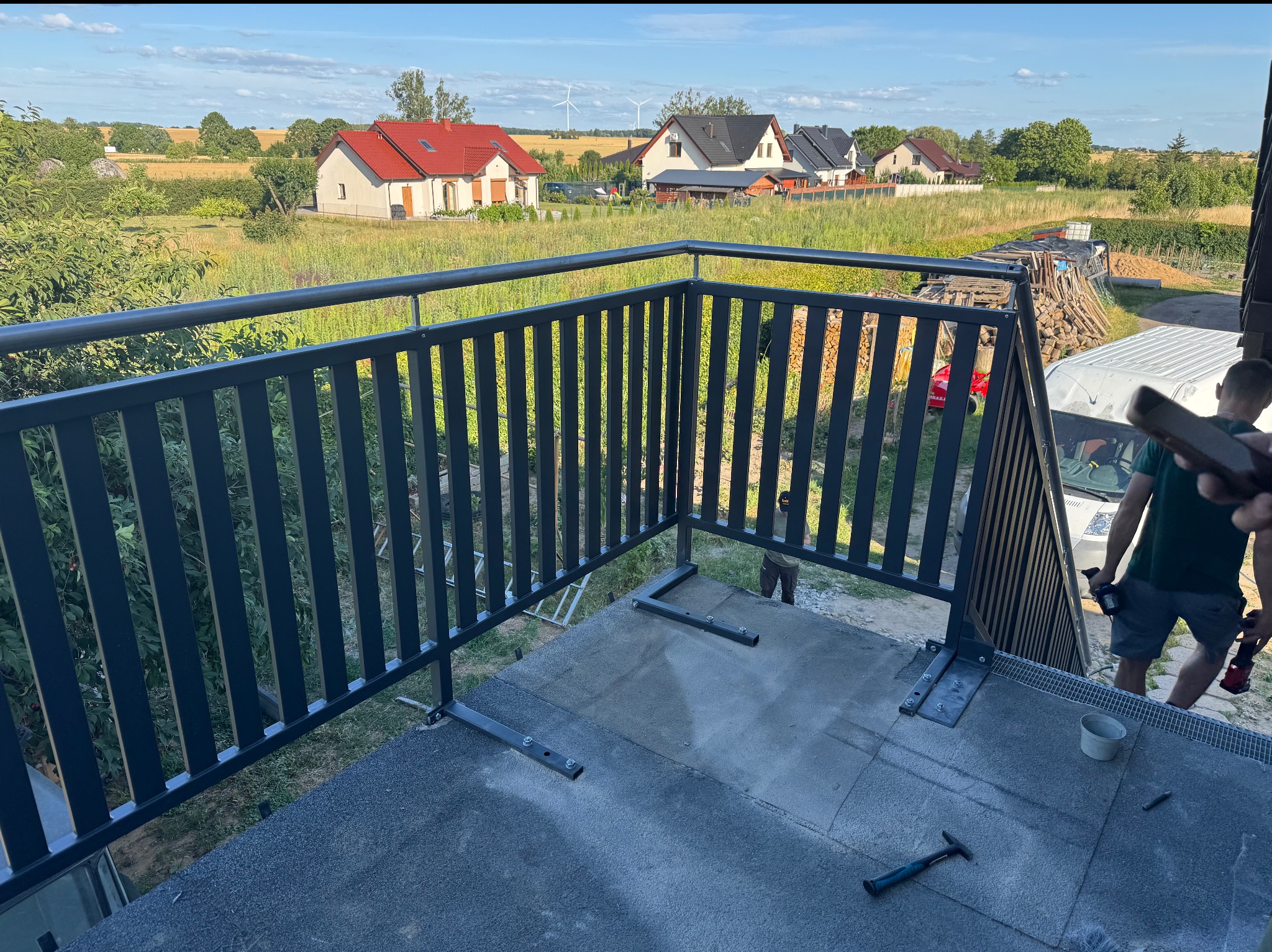 Montaż metalowej balustrady balkonowej z pionowymi szczeblami, widok na okoliczne domy i pola w słoneczny dzień. Etap mocowania balustrady do podłoża.