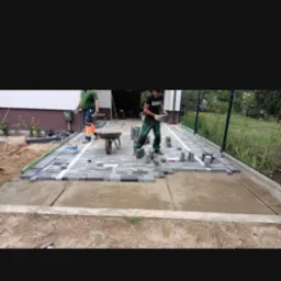 Dw&oacute;ch pracownik&oacute;w układa szarą kostkę brukową na przygotowanym podłożu. Jeden z nich trzyma kostkę w rękach, drugi pracuje z taczką. Częściowo ułożona nawierzchnia z widoczną białą linią...