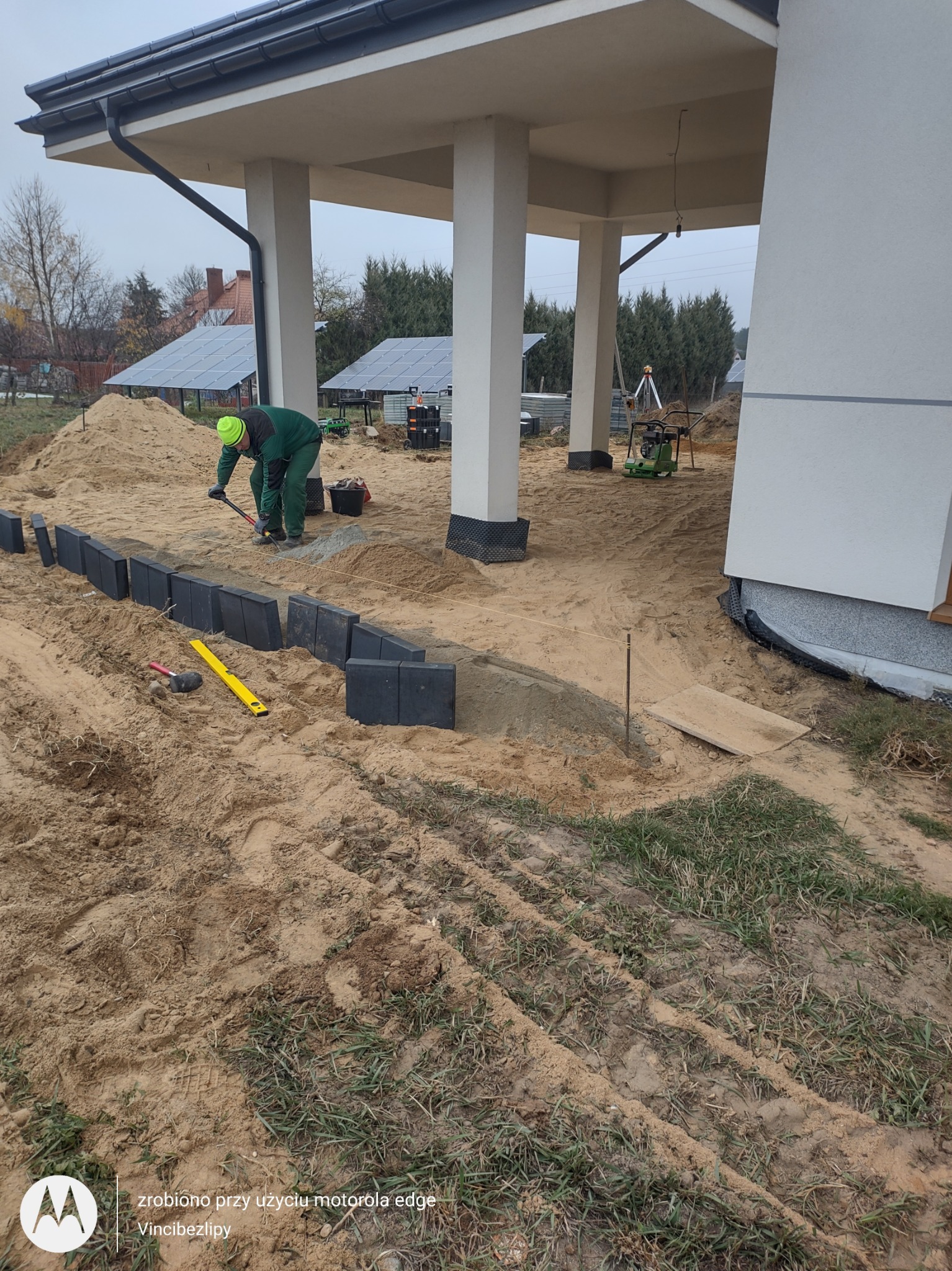 Pracownik w zielonym stroju układa czarną kostkę brukową wzdłuż fundamentów domu, obok leżą poziomica i gumowy młotek, w tle widać panele solarne.