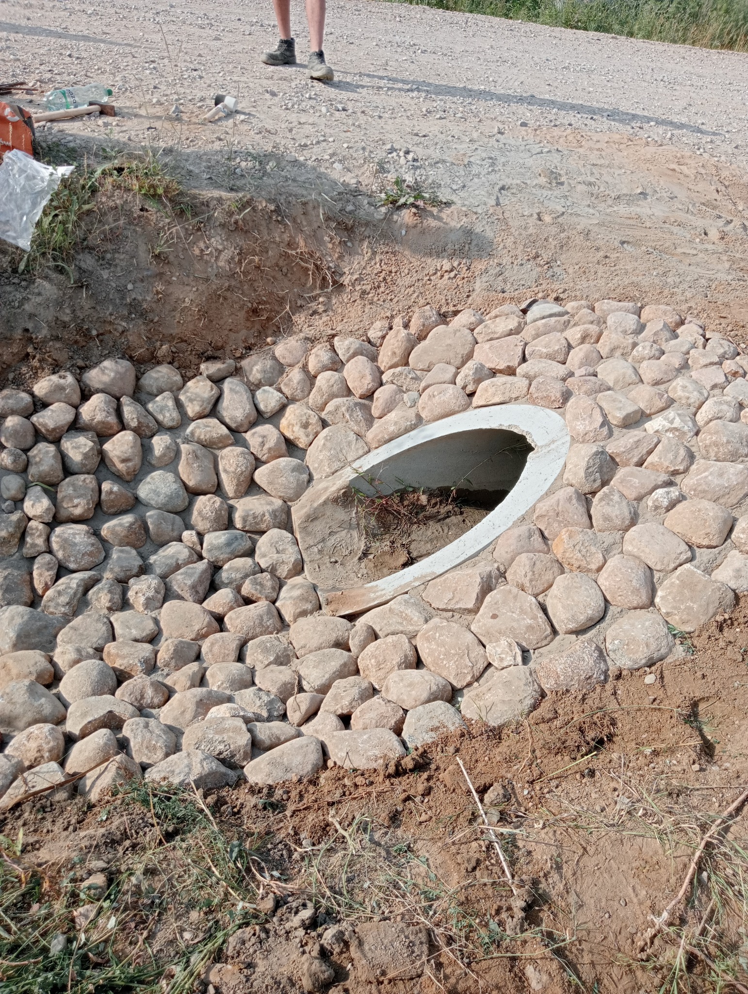 Przepust drogowy z betonową rurą otoczony ułożonymi kamieniami rzecznymi, widoczne ślady ziemi i trawy.