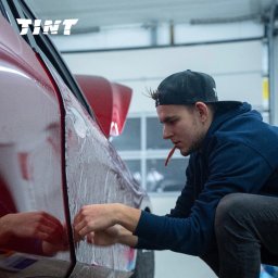 TINT Oklejanie Pojazdów Michał Olczyk - Mężczyzna w czapce aplikuje folię na czerwony samochód. Widoczny bok auta, narzędzia w ustach aplikatora. Logo firmy w górnym rogu.
