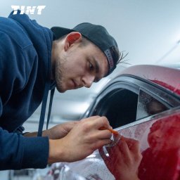 TINT Oklejanie Pojazdów Michał Olczyk - Młody mężczyzna w czapce aplikuje folię ochronną na szybę czerwonego samochodu, używając pomarańczowego narzędzia. Widoczne logo firmy.