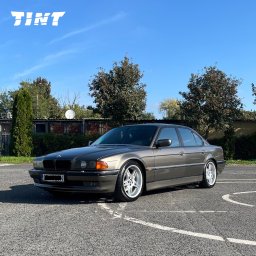 TINT Oklejanie Pojazdów Michał Olczyk - Szary, luksusowy sedan BMW serii 7 (E38) z przyciemnionymi szybami bocznymi, zaparkowany na parkingu w słoneczny dzień. Widoczne logo firmy 'TINT' w lewym górnym rogu.