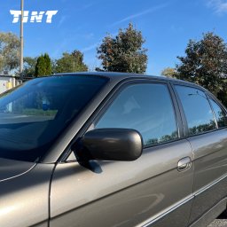 TINT Oklejanie Pojazdów Michał Olczyk - Ciemne szyby w aucie marki BMW, widok z boku, na tle błękitnego nieba i drzew. Usługa wykonana profesjonalnie.