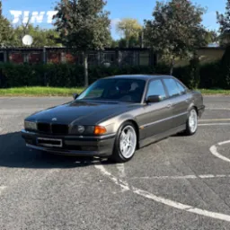 Szary BMW serii 7 (E38) z aluminiowymi felgami na parkingu w słoneczny dzień. Widoczne logo firmy na szybie, sugerujące przyciemnianie szyb.