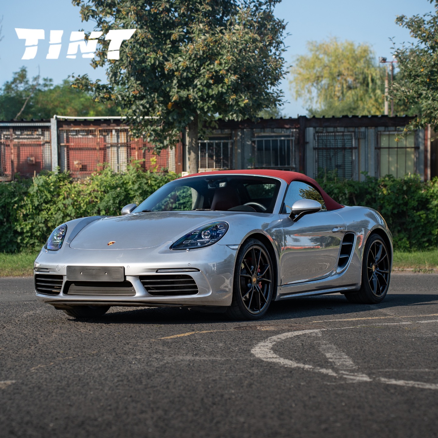 Srebrny Porsche Boxster z czerwonym dachem na tle zieleni i metalowych konstrukcji, logo firmy w górnym rogu, wyraźne odbicia w lakierze.