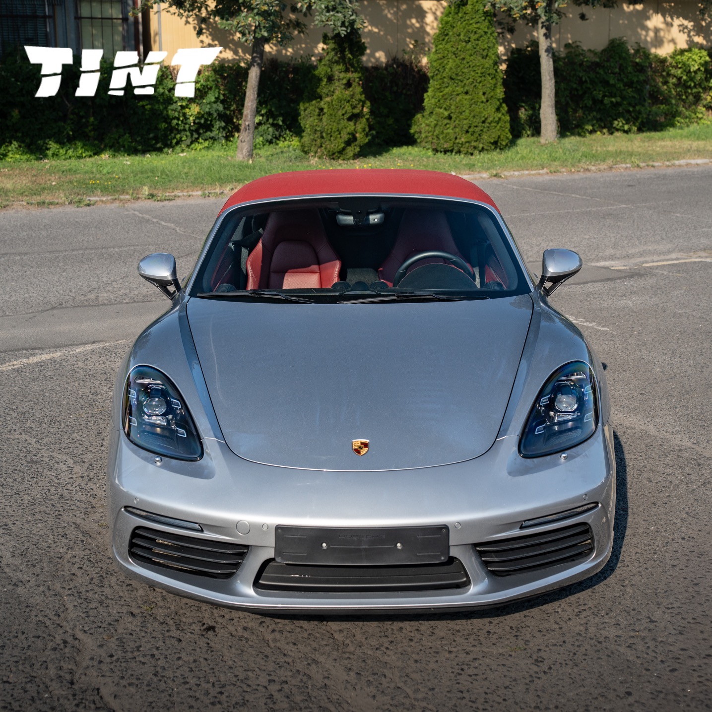 Srebrny Porsche 718 z czerwonym dachem kabrioletu, widziany z przodu na zewnątrz. Logo firmy w lewym górnym rogu.